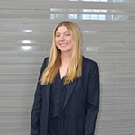 Professional portrait of a woman with long blonde hair, wearing a dark blazer and standing in front of a modern background.