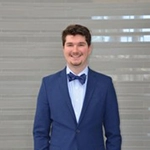 A young man in a formal blue suit and bow tie stands smiling against a gray background.