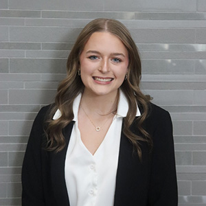 Woman with long brown hair, smiling and wearing a white shirt and dark jacket.