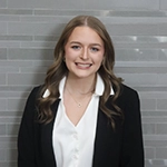 A young woman with long hair wearing a black blazer over a white blouse, smiling against a gray tiled background.