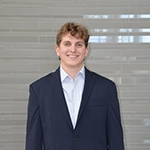 A young man in a professional suit and shirt stands smiling against a modern backdrop.