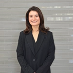 Professional headshot of a woman in a black blazer, smiling in front of a light gray background.