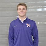 Portrait of a young man wearing a purple Nike jacket with a logo on the left chest, smiling against a light gray background.