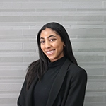 Professional portrait of a young woman smiling, wearing a black blazer against a minimalist gray background.