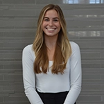 Portrait of a smiling woman with long, wavy hair wearing a white top, standing in front of a gray tiled background.