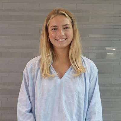 A young woman with long blonde hair wearing a light blue button-up shirt, smiling in front of a gray textured wall.