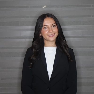 Professional headshot of a smiling woman in a black blazer against a gray tiled background.