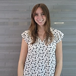 Smiling young woman in a patterned blouse stands in front of a gray wall.