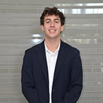 A young man in a navy blazer and white shirt stands smiling in front of a light-colored wall.