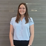 A smiling young woman in a light blue blouse poses in front of a gray wall.