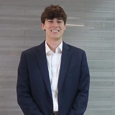 A young man wearing a navy suit jacket and white dress shirt, smiling at the camera against a light gray textured background.