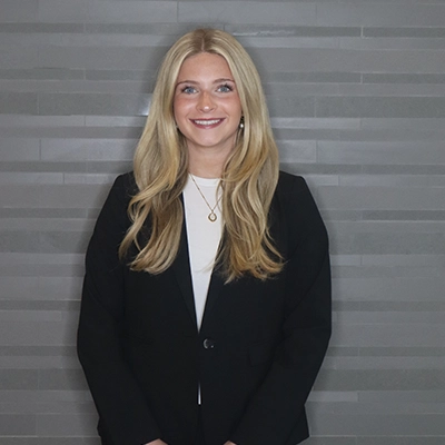 Professional headshot of a young woman in a black blazer with long blonde hair, standing against a gray tiled background.