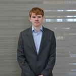 A young man in a formal suit stands against a neutral backdrop.