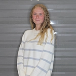 A smiling young woman with long, wavy hair stands against a gray backdrop, wearing a light-colored striped sweater.
