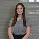 Professional headshot of a young woman in a gray blouse, smiling with arms crossed, set against a softly blurred background.