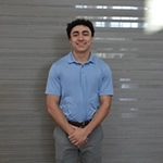 A young man in a light blue shirt and gray pants stands with his hands clasped in front of a neutral background.