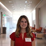 Young woman with brown hair smiling in a brightly lit hallway, wearing a red blouse and name tag.