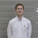 Smiling man in a light checkered button-down shirt standing in front of a gray, horizontally striped wall.
