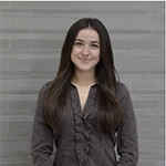 Portrait of a smiling woman with long dark hair wearing a gray blouse, standing in front of a light gray brick wall.