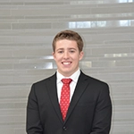 A professional headshot of a young man in a suit and tie, smiling against a light, modern background.