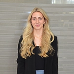 Professional headshot of a woman with long blonde hair wearing a black blazer and standing against a neutral background.