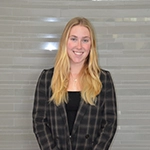 Professional headshot of a woman with long blonde hair, wearing a black and white checked blazer and smiling confidently.