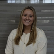 Smiling woman with long blonde hair wearing a light-colored button-up shirt, seated indoors in front of a gray tiled wall.
