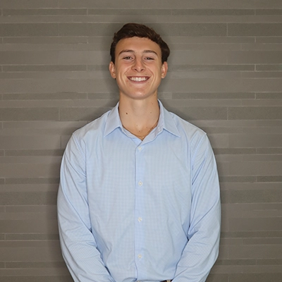 A smiling young man in a light blue dress shirt stands against a textured gray wall.