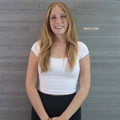 A smiling young woman with long blonde hair standing against a textured gray background.