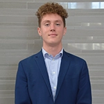 A young man with curly hair wearing a blue suit and light blue shirt, standing against a textured background.