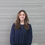 A smiling woman with long brown hair wearing a dark blue dress stands in front of a light gray wall.