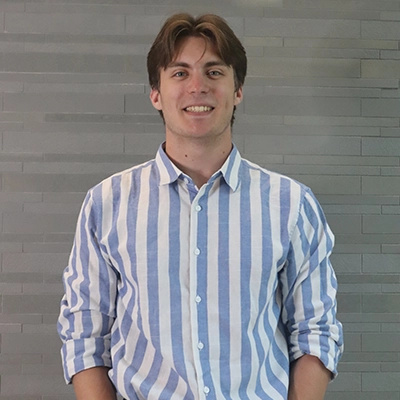 A young man smiling in a casual blue and white striped shirt against a gray textured background.