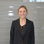 A young woman in a black blazer stands smiling against a light gray background.