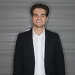 Smiling young man in a suit and white shirt standing against a gray background.