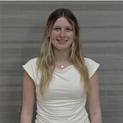Smiling young woman with long blonde hair wearing a white top, standing in front of a gray tiled wall.