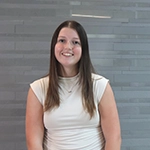 A smiling woman with long brown hair is posing in front of a gray textured wall.