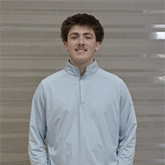 Person in a light blue quarter-zip pullover standing indoors against a beige tiled wall.