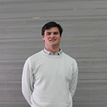 A young man smiling, standing against a gray background, wearing a white sweater and collared shirt.