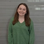A smiling young woman wearing a green sweater stands against a light gray background.
