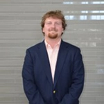 Professional headshot of a man in a suit and tie, standing confidently against a neutral background.
