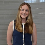 A young woman with long hair smiles confidently while wearing a navy dress with white buttons.