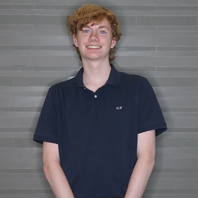 A young man with curly red hair smiles while wearing a navy polo shirt.