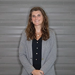 A smiling woman with long brown hair stands in front of a gray wall, wearing a black top and a gray cardigan.
