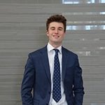 A professional young man in a suit and tie, standing against a modern background.