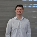 Smiling young man wearing a white shirt, standing in front of a gray wall with horizontal lines.
