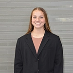 A smiling young woman wearing a black blazer and light brown top, standing in front of a gray backdrop.
