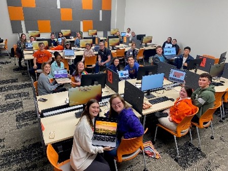 A group of students in a computer lab holding up laptops, showcasing their work and engaged in a collaborative learning environment.