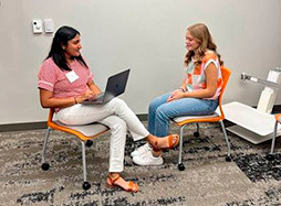 Two young women engage in a conversation, one using a laptop while sitting on orange chairs in a modern office setting.