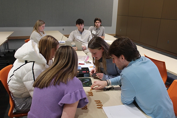 Group of students collaborating around a table in a classroom, working on a hands-on project with paper and markers.
