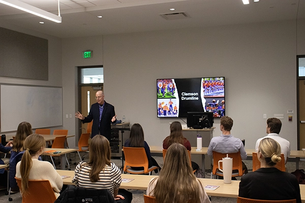 Presenter speaking to an audience in a classroom, with a screen displaying a slide titled Clemson Drumline.
